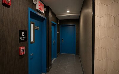 Dark contemporary hotel corridor with blue guest and exit doors, gray tiled flooring, recessed ceiling lights, and clearly marked fire and exit signage.