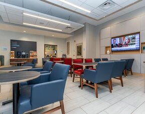 Contemporary hotel dining and lounge area with blue and red seating, round tables, beverage station, wall-mounted televisions, and modern lighting.