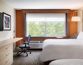 Bright, modern hotel room with two white beds, a work desk and chair, wall-mounted TV, and large window overlooking green trees.