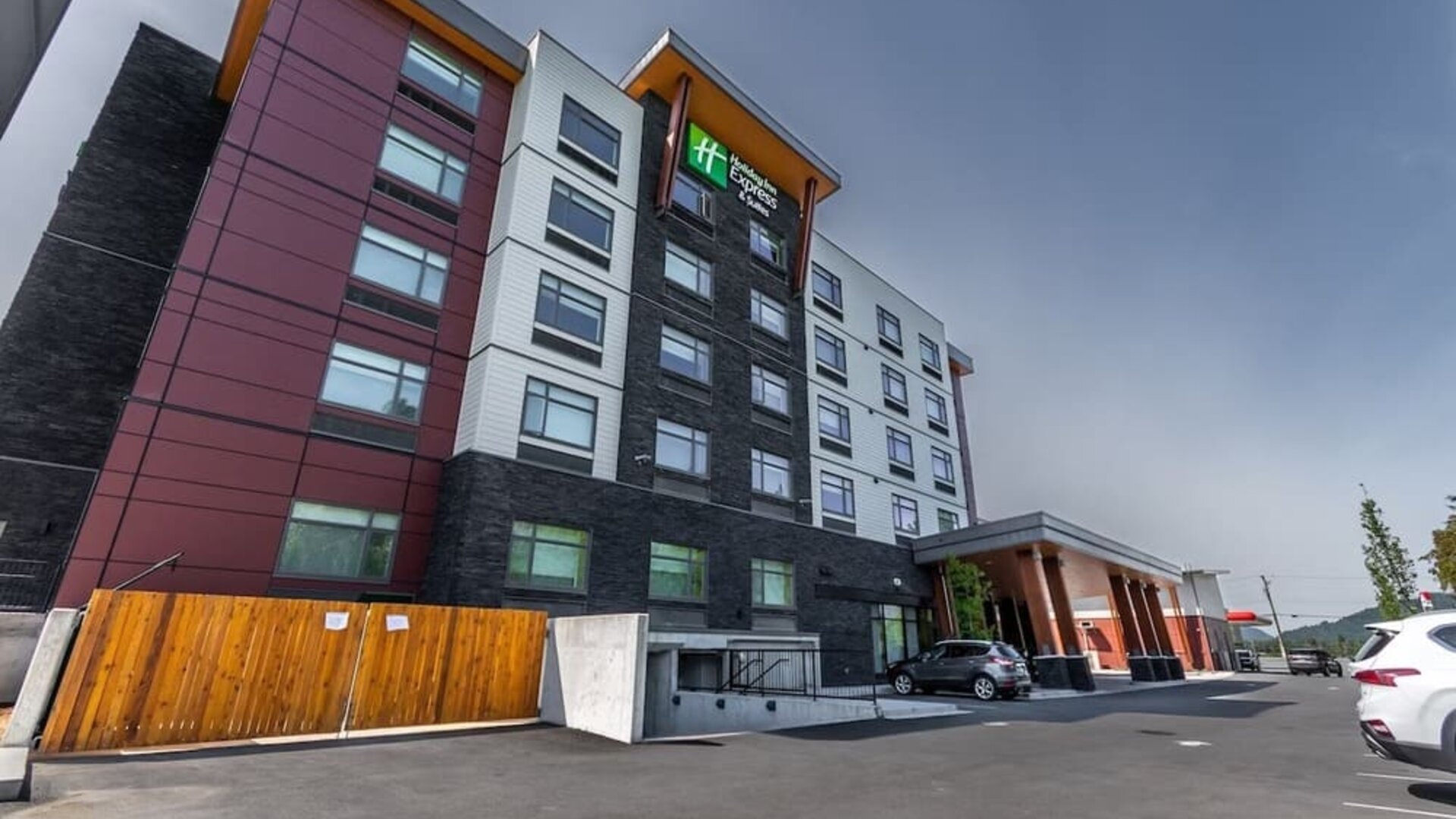 Exterior view of a modern Holiday Inn Express hotel with multi-story red, white, and dark stone façade, covered entrance, and adjacent parking area.