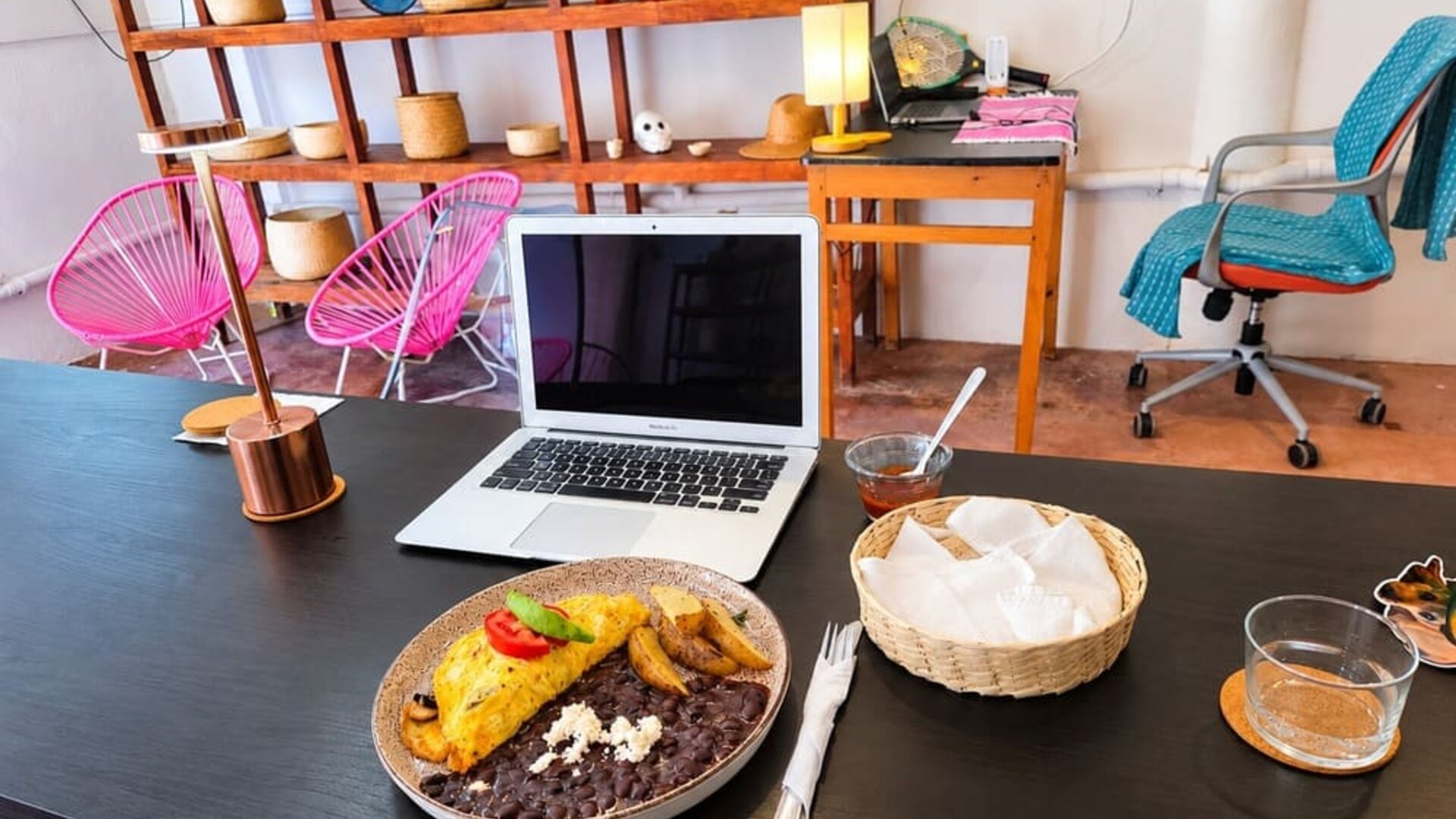 Open laptop beside a plated breakfast of omelet, black beans, potatoes, and cheese on a spacious coworking table with colorful seating and decorative shelving.