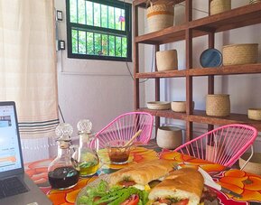 Sandwich and green salad served beside a laptop on a vibrant floral table, with condiment bottles, pink woven chairs, wooden shelving, and natural light.