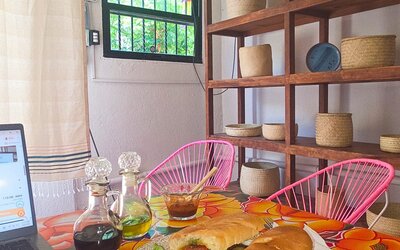 Sandwich and green salad served beside a laptop on a vibrant floral table, with condiment bottles, pink woven chairs, wooden shelving, and natural light.