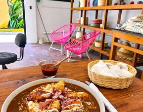 Plate of Mexican-style food with fried plantains, sauce, cheese and toppings served on a table with salsa and napkins, surrounded by wooden shelving, woven décor, bright pink chairs, and a window overlooking tropical greenery.
