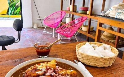 Plate of Mexican-style food with fried plantains, sauce, cheese and toppings served on a table with salsa and napkins, surrounded by wooden shelving, woven décor, bright pink chairs, and a window overlooking tropical greenery.