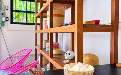 Colorful coworking café interior with a large plate of Mexican-style food and fried plantains on a wooden table, salsa and napkins, bright pink woven chairs, wooden shelving, and tropical greenery visible outside.
