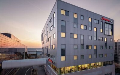 Modern Hampton hotel exterior at sunset with a gray multi-story façade, illuminated guest room windows and an enclosed walkway connecting to a neighboring building.