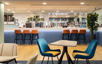 Contemporary hotel bar and lounge with a long blue counter, orange bar stools, pendant lighting, stocked drinks display, wooden flooring and blue lounge chairs.