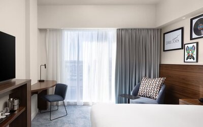 Bright modern hotel room with a large bed, floor-to-ceiling window and curtains, blue armchair, wooden work desk, chair, wall-mounted TV and contemporary artwork.