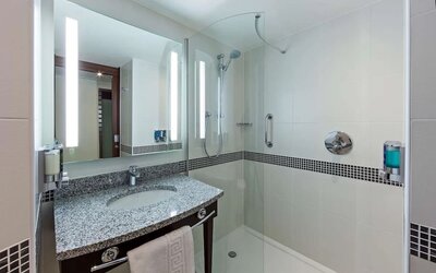 Modern hotel bathroom with a granite vanity, illuminated mirror, glass-enclosed walk-in shower, handheld showerhead, grab rail and wall-mounted toiletries.