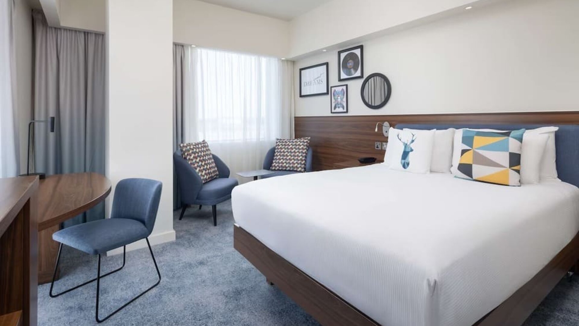 Spacious modern king room with a large white bed, blue upholstered headboard, colorful decorative pillows, two armchairs, work desk and large curtained windows.