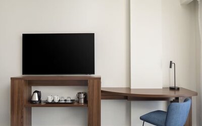 Hotel room workspace with a large flat-screen TV, curved wooden desk, blue chair, desk lamp and hospitality station with kettle, cups, glasses and ice bucket.