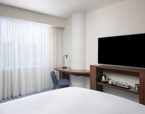 Bright contemporary hotel room with a large bed, floor-to-ceiling sheer curtains, wooden work desk, blue chair, wall-mounted TV and tea and coffee station.