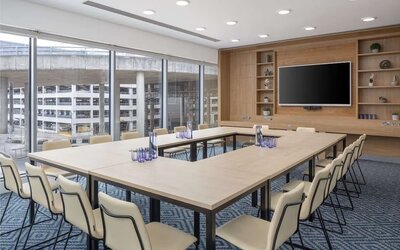 Bright hotel meeting room with U-shaped conference tables, upholstered chairs, large wall-mounted TV, wooden shelving and floor-to-ceiling windows overlooking nearby buildings.