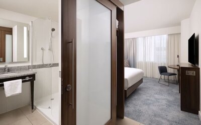 Contemporary hotel room viewed from the entrance, with a king bed, work desk and TV beside a modern bathroom with a glass-enclosed shower and illuminated mirror.