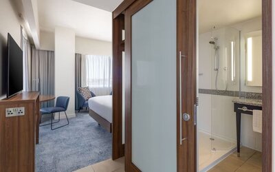 Modern hotel king suite with a bedroom and bathroom separated by a frosted-glass sliding door, featuring a work desk, wall-mounted TV and glass-enclosed shower.