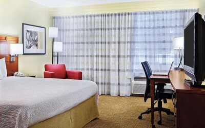 Spacious hotel king room with white bedding, wooden headboard, red sofa, coffee tables, work desk, large curtained window and warm yellow walls.