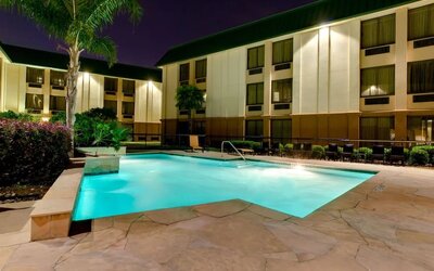 Illuminated outdoor hotel swimming pool surrounded by stone decking, lounge chairs, tropical landscaping and multi-story guest room buildings at night.
