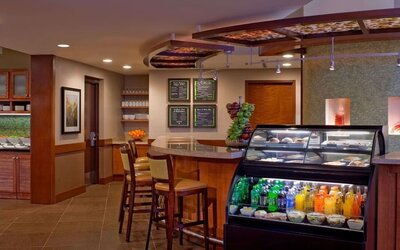 Casual hotel café and dining area with a refrigerated display case stocked with drinks and snacks, counter seating, wooden cabinetry, menu boards, and tables for guests.