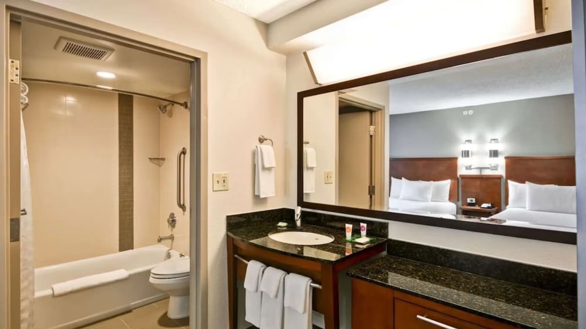 Modern hotel guest bathroom with a bathtub and shower, granite vanity with sink, large framed mirror, fresh white towels, and a view into a guest room with two beds.