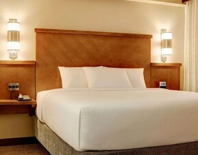Comfortable hotel king bed dressed in crisp white linens and pillows, framed by a warm wooden headboard, bedside tables, contemporary wall lights, and a large window with curtains.