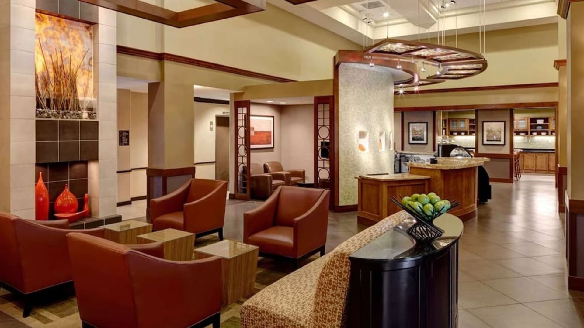 Stylish hotel lobby lounge with rust-colored armchairs, small side tables, contemporary artwork, warm wood accents, tiled flooring, and an open café area in the background.