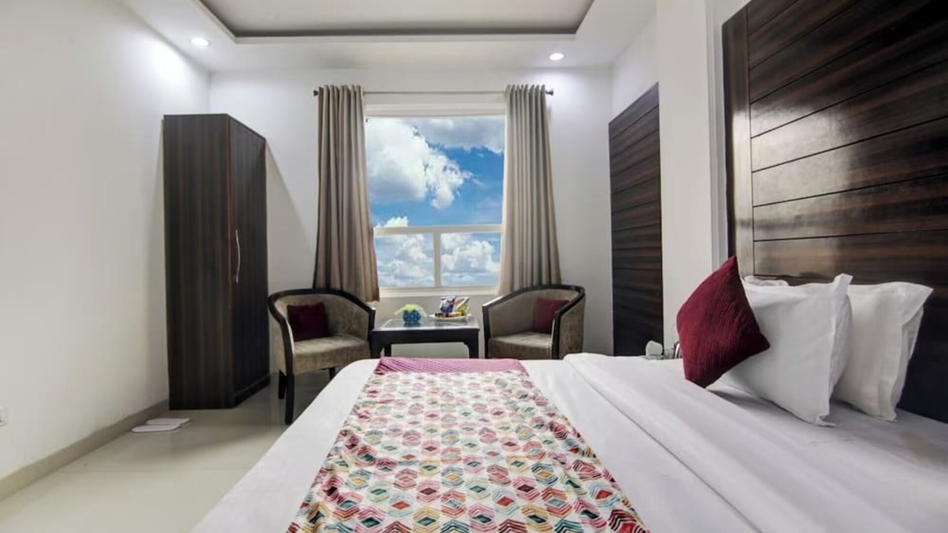 Bright hotel guest room with a neatly made bed featuring white linens, burgundy accent pillows and a colorful patterned runner, plus a wall-mounted TV, wardrobe, two armchairs, small table, ceiling fan and large window.