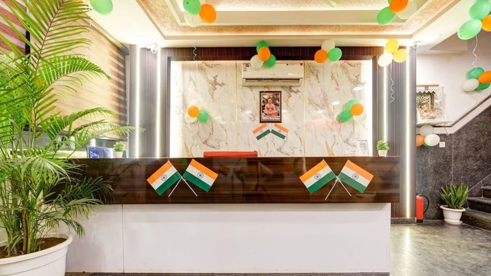 Hotel reception area decorated with Indian tricolor balloons and flags, featuring a dark wood and white front desk, marble-patterned feature wall, indoor plants and staircase to the side.