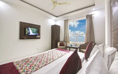 Modern hotel room with a large bed dressed in white linens and red patterned accents, dark wood feature headboard, ceiling fan, large window with curtains, wall-mounted TV, seating area and small glass-topped table.