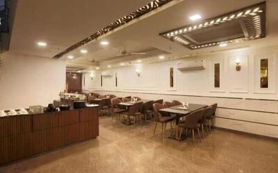 Spacious hotel dining area with multiple tables and upholstered chairs, buffet counter, polished flooring, decorative ceiling panels, recessed lighting, ceiling fans and wall-mounted air-conditioning units.
