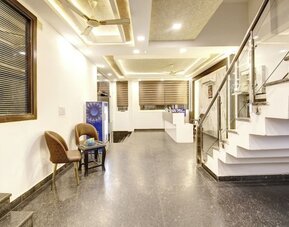 Bright interior hotel entrance and lobby corridor with polished stone flooring, two chairs and a small table, illuminated ceiling details, glass-railed staircase and reception area visible in the background.