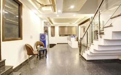 Bright interior hotel entrance and lobby corridor with polished stone flooring, two chairs and a small table, illuminated ceiling details, glass-railed staircase and reception area visible in the background.