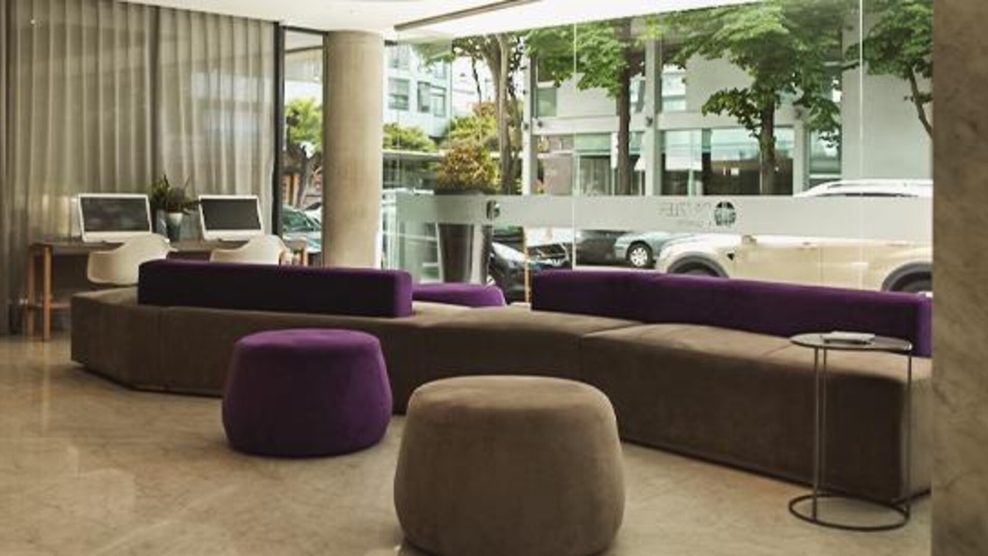 Hotel lobby with purple and gray sofas, round ottomans, floor-to-ceiling windows, and street views at Dazzler by Wyndham Polo.
