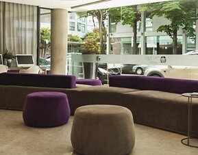 Hotel lobby with purple and gray sofas, round ottomans, floor-to-ceiling windows, and street views at Dazzler by Wyndham Polo.