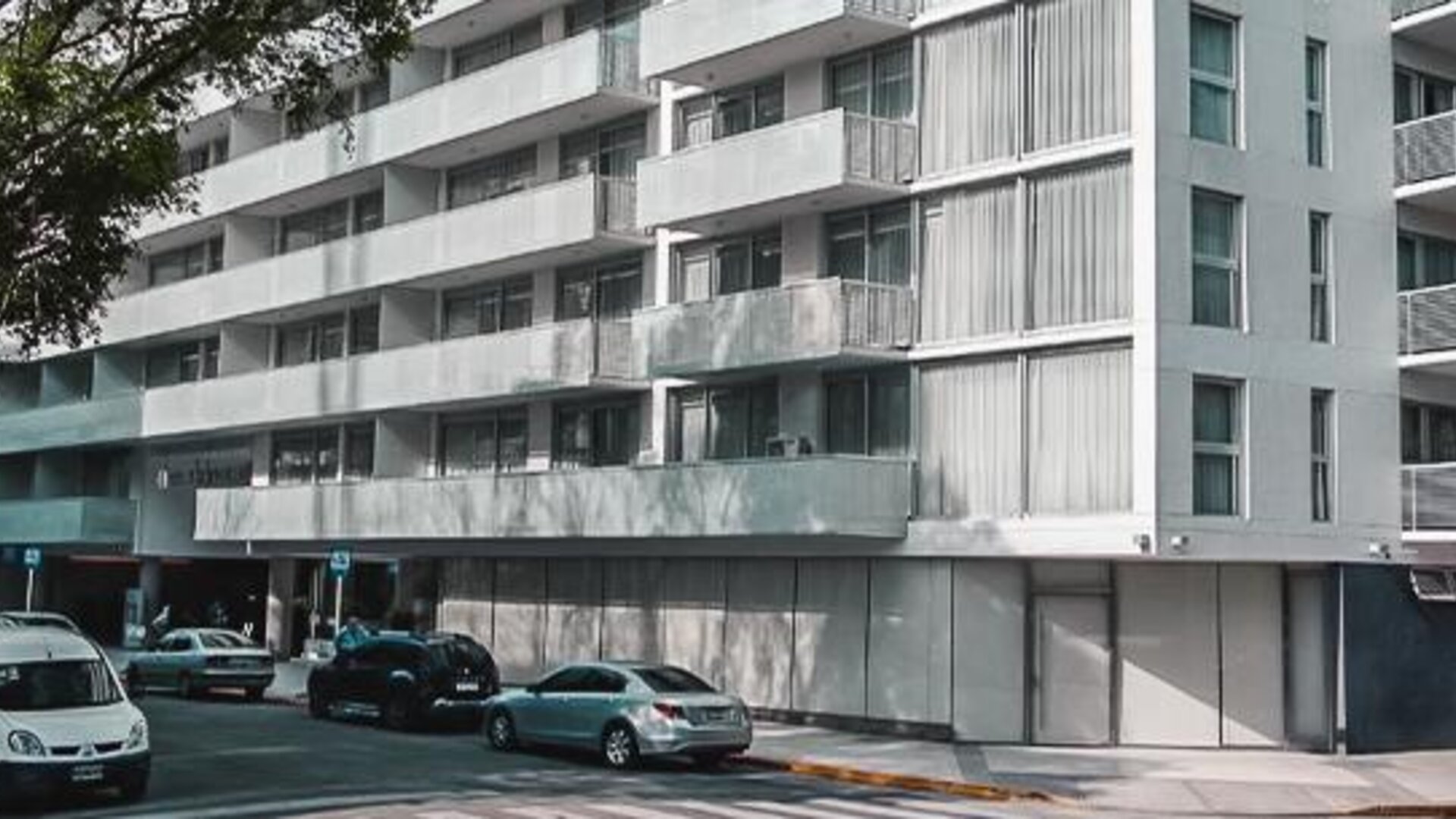 Modern multi-story hotel exterior with balconies, street-level windows, trees, and parked cars at Dazzler by Wyndham Polo.