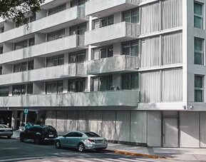 Modern multi-story hotel exterior with balconies, street-level windows, trees, and parked cars at Dazzler by Wyndham Polo.
