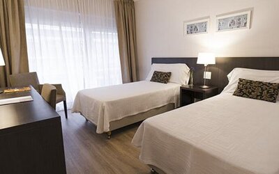 Twin-bed guest room with a desk, television, armchair, bedside table, and large curtained window at El Conquistador Hotel.