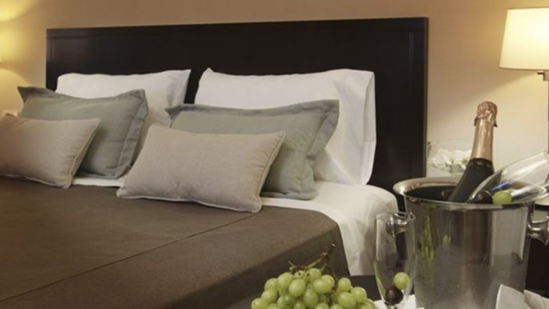 Close view of a king bed with layered pillows, dark headboard, bedside lamps, and champagne at El Conquistador Hotel.