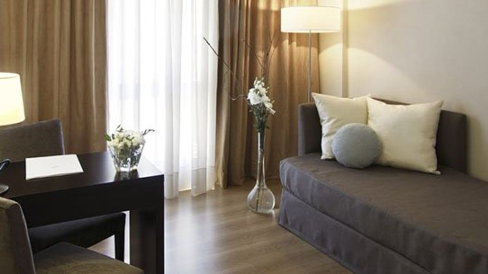 Guest room with a single sofa bed, desk and chair, floor lamp, and curtains beside a bright window at El Conquistador Hotel.