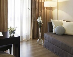 Guest room with a single sofa bed, desk and chair, floor lamp, and curtains beside a bright window at El Conquistador Hotel.