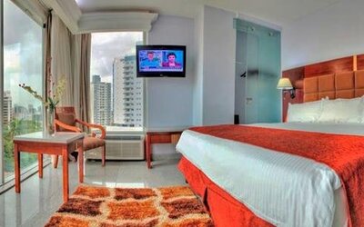 Guest room with a king bed, red runner, television, desk, chair, and floor-to-ceiling windows with city views at Hotel Grand International.