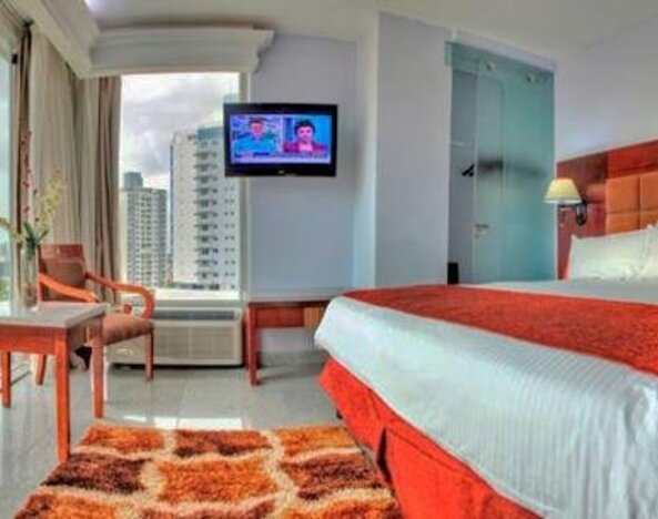 Guest room with a king bed, red runner, television, desk, chair, and floor-to-ceiling windows with city views at Hotel Grand International.
