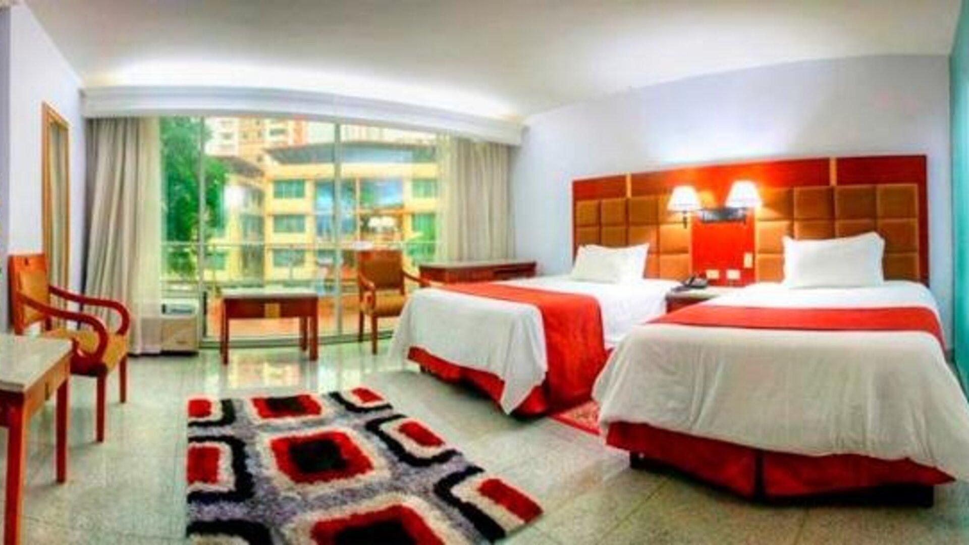 Guest room with two beds, red runners, seating area, patterned rug, and large windows overlooking the surroundings at Hotel Grand International.