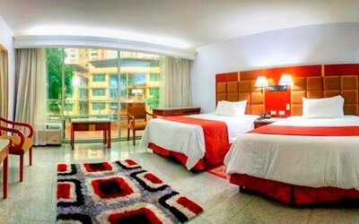 Guest room with two beds, red runners, seating area, patterned rug, and large windows overlooking the surroundings at Hotel Grand International.