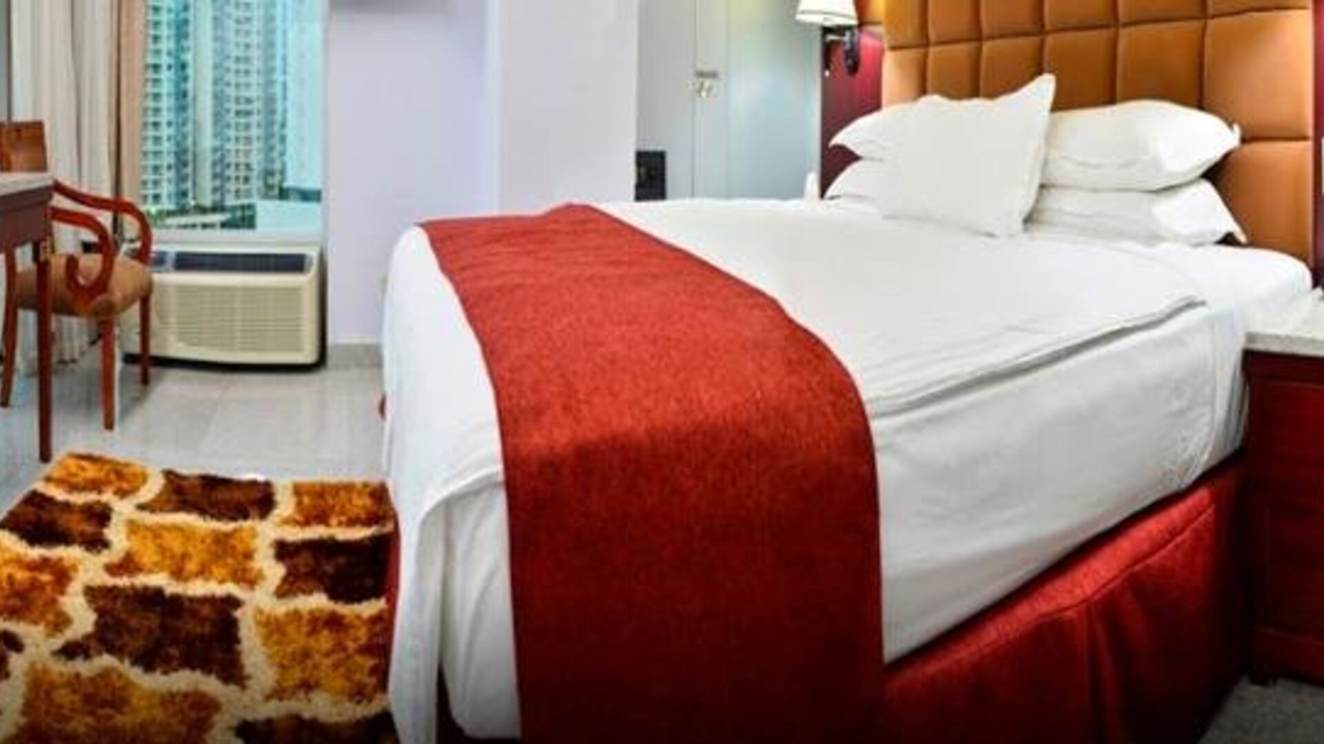 Guest room with a king bed, red runner, wood furnishings, desk, and large windows overlooking city buildings at Hotel Grand International.