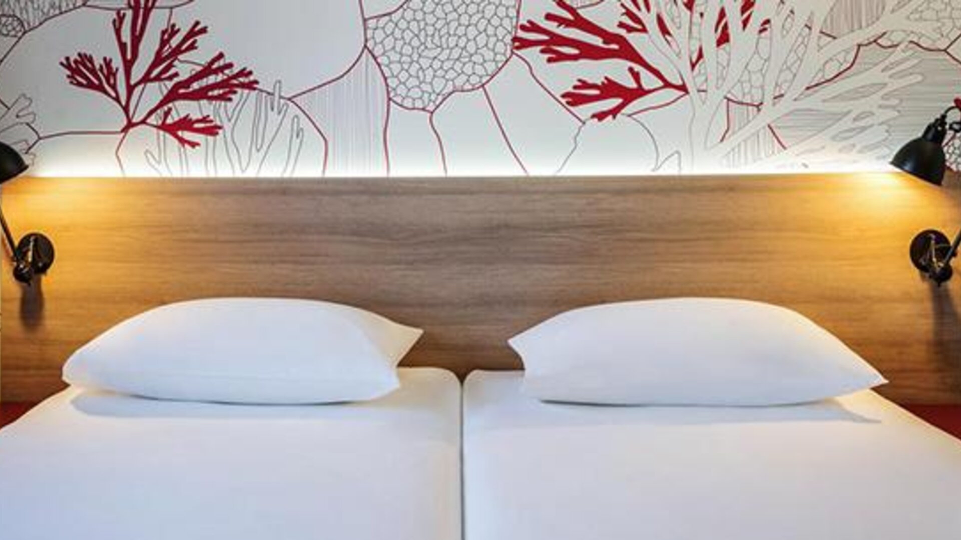 Close view of twin beds with white linens, wooden headboard, reading lamps, and coral-patterned wall at Hotel ibis Styles Barcelona City Bogatell.
