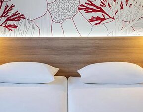 Close view of twin beds with white linens, wooden headboard, reading lamps, and coral-patterned wall at Hotel ibis Styles Barcelona City Bogatell.