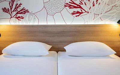 Close view of twin beds with white linens, wooden headboard, reading lamps, and coral-patterned wall at Hotel ibis Styles Barcelona City Bogatell.