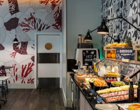 Dining area with rows of tables and chairs, pendant lights, sofas, and patterned decorative walls at Hotel ibis Styles Barcelona City Bogatell.