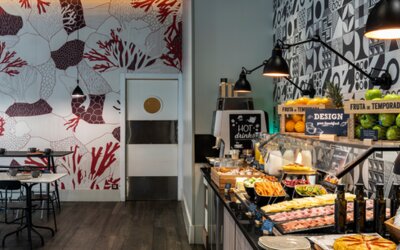 Dining area with rows of tables and chairs, pendant lights, sofas, and patterned decorative walls at Hotel ibis Styles Barcelona City Bogatell.